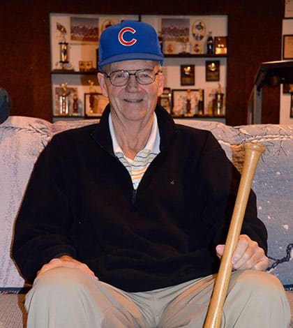 Nelson Mathews sits on his couch in Columbia while wearing a Cubs hat and holding a bat used during his playing days.